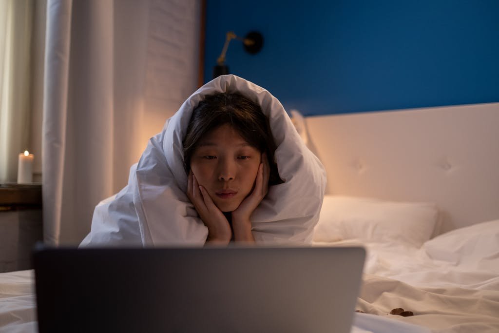 Woman bundled in blankets watching a laptop screen in a cozy bedroom setting.